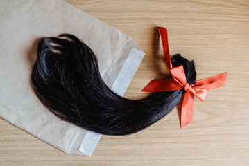 Mechón de pelo cortado anudado con un lazo de raso rojo metido en un sobre de color marrón. Enviar una donación de cabello para pelucas para pacientes con cáncer mediante carta - obrazy, fototapety, plakaty