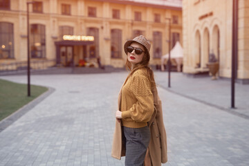 Turned from behind fashion model young woman in sunglasses and hanging on a shoulder autumn beige coat walking on the street happily posing for the camera. Tinted photo. 