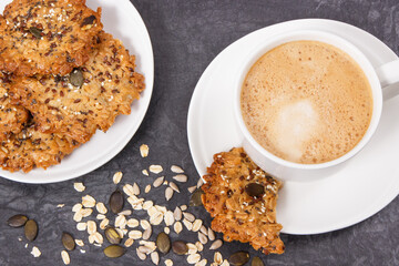 Cup of coffee with milk and fresh oatmeal cookies with honey and healthy seeds on white plate. Delicious crunchy dessert