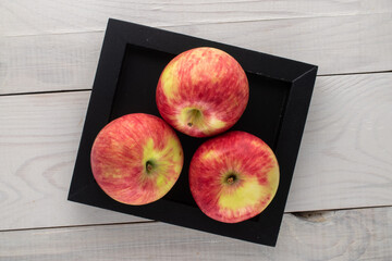 Three juicy apples in a black tray made of wood on a wooden table, top view.