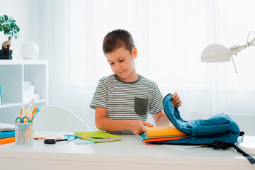 schoolboy putting school stationery into backpack at table indoors at home white room.