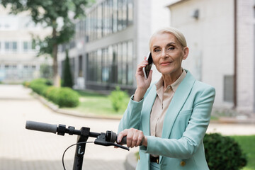Fototapeta premium elderly businesswoman talking on mobile phone near electric kick scooter outdoors