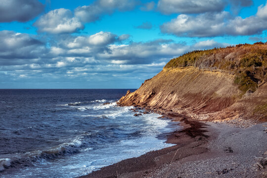 Evening is approaching the rugged coast of Cape Breton. The sun shines upon the face of a cliff while waves of the cool waters of the Atlantic Ocean land upon the rocky beach.
