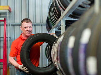 Car service. Male worker checking new tire wheel on shelves shelf at wheel store, wearing red uniform, gloves during working. Specialist technician worker examining new tire wheel. Maintenance concept