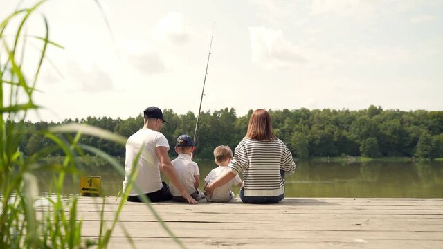 young happy family with little boy fishing a camping site. Parents with kids fishing together sitting on a wooden pier by pond spending weekend holiday outdoors. Back Rear view. catching the fish