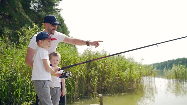 father with two little children sons catches fish standing on a wooden bridge or pier. Family dad and kids spend leisure time together camping teach fishing. Day vacation on lake or river outdoors