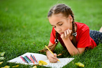 child drawing on a grass A girl in a red t-shirt lying on the green grass with yellow leaves in the park and drawing with colored pencils