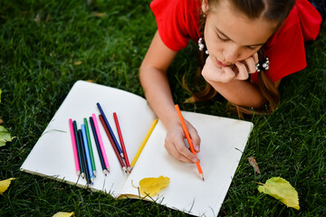 child drawing on a grass A girl in a red t-shirt lying on the green grass with yellow leaves in the...
