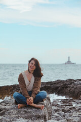 Fototapeta na wymiar woman sitting by rocky sea beach in wet jeans lighthouse on background