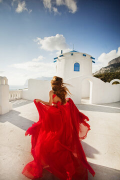 Fashion outdoor photo of gorgeous young woman with red hair in airy red dress posing on the streets of Oia, Santorini, Greece. 