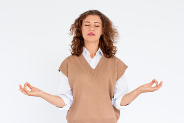 Curly beauiful young female makes mudra sign, relaxes after hard working day, keeps eyes shut,...