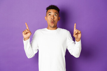 Image of impressed african-american guy in white sweatshirt, pointing and looking up with amazed...