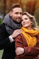 Tenderness of a cute couple. Handsome man and a woman hugged from behind smile looking at each other in the autumn park. Outdoor shot of a young couple in love having great time.