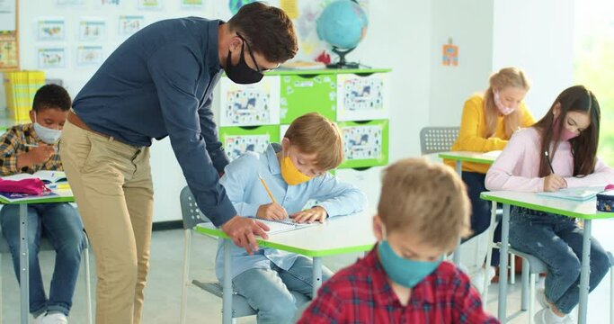Caucasian young male teacher in mask helping small junior student studying in classroom. Elementary School. Mixed-race children learning after coronavirus lockdown. Lesson, primary school education
