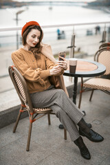 Fototapeta na wymiar Charming french young woman using her smartphone taking selfie while having cup of coffee. Frenchwoman wearing red beret sitting on terrace of restaurant or cafe with background of autumn urban city. 