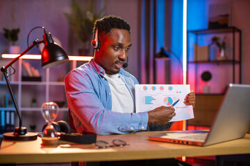 African american man in headset having video conference on modern laptop. Young guy in casual wear sitting showing financial report during working meeting online. Remote work concept.