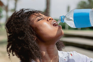 Afro woman drinking water from a bottle while walking outdoors on a hot and sunny day.