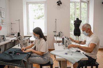 Multiethnic tailor and seamstress in medical masks working with cloth near sewing machine