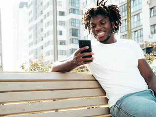 Handsome hipster model.Unshaven African man dressed in white summer t-shirt.Fashion male with dreadlocks hairstyle sitting at the bench in the street.Looking at smartphone screen, using cellphone apps