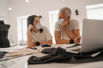 Obraz premium Interracial designers in medical masks talking near laptop and cloth