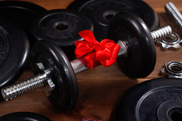 Black dumbbell with a red bow for a gift on a wooden table near sports equipment. A gift to the athlete. Healthy lifestyle, sport