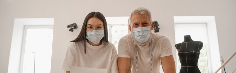 Interracial designers in medical masks looking at camera in atelier, banner