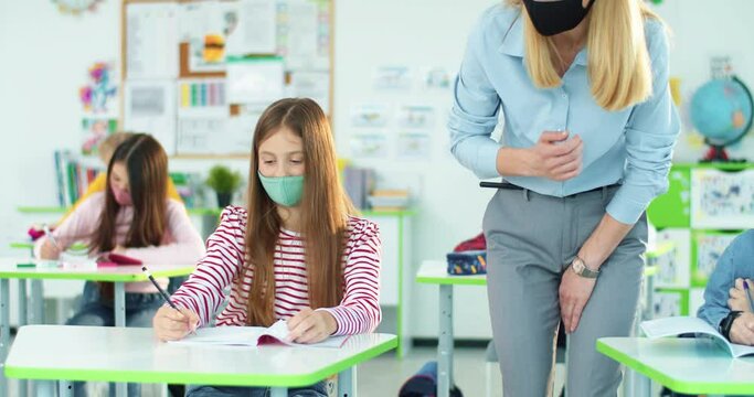 Portrait of Caucasian young beautiful woman teacher in mask teaching class and explaining pupils school material helping to write a test. Children studying in classroom writing exam. Education concept