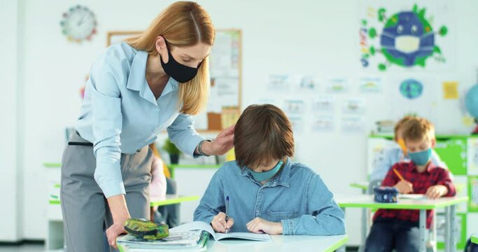 Portrait of beautiful Caucasian woman tutor helping little junior student to write test in copybook at classroom. Female teacher explaining to smart schoolboy lesson task. Post-covid education