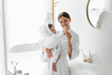 happy mother holding in arms toddler son in bathrobe and toothbrush