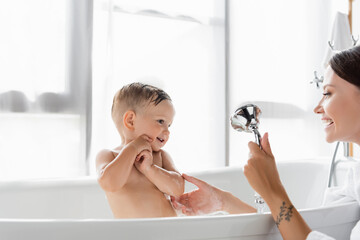 cheerful mother with tattoo holding shower head while bathing toddler boy in bathtub