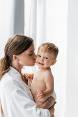 tattooed mother in bathrobe holding in arms and kissing cheek of nude toddler son