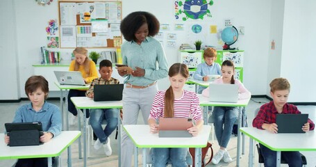 Multi-ethnic diverse junior students boys and girls studying at school computer technology lesson using laptops and tablets computer science class African American young female teacher teaching pupils - Powered by Adobe