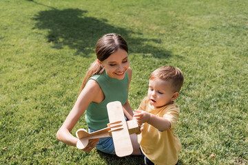 smiling mother holding toy biplane near toddler son on grass