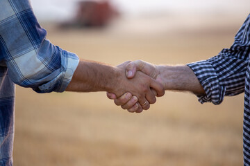 Two farmers in plaid shirt shaking hands in front of combine harvester working in wheat field in summer time. Agribusiness and making deals concept