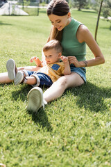 Naklejka na ściany i meble cheerful mother sitting on grass with toddler son outside