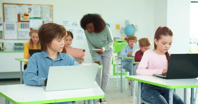 Children learning at primary school. Portrait of Caucasian schoolgirl and schoolboy typing on laptop surfing internet at computer science class African American female teacher tapping on tablet behind