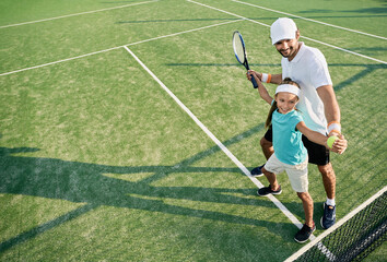 Positive little girl while learning to play tennis with male coach on outdoor grass court....