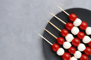 Cold appetizer on wooden skewers: red cherry tomatoes and balls of mozzarella cheese on a black plate against a gray stone background. Copy space
