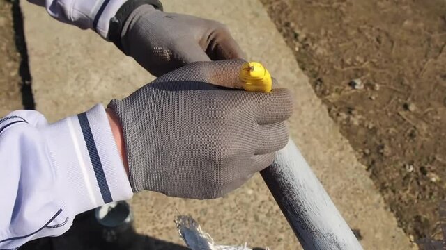 Person paints pipe with primer outdoors. Close up hand in gloves which hold yellow brush and cover iron tube. Home improvement and DIY concept. Protection of metal square pipes from rust and corrosion