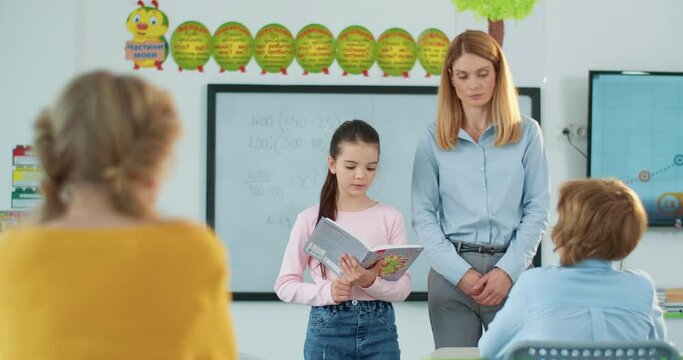 Little cute Caucasian schoolgirl answering question reading from textbook while standing in the middle of classroom in front of classmates and pretty female teacher. Studying, elementary education