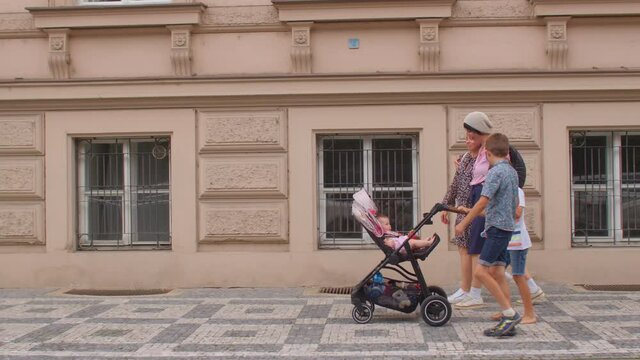 Young women with a stroller and children walk along a deserted street in a European city. Facades with windows and doors of houses of the old town of Prague. Walking with kids.