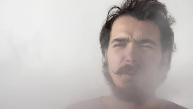 Man is covered by cigarette fume done by somebody who consumes tobacco next to him. Isolated portrait of a male person suffering from passive smoking. He is coughing and waving hand. Harm of nicotine.