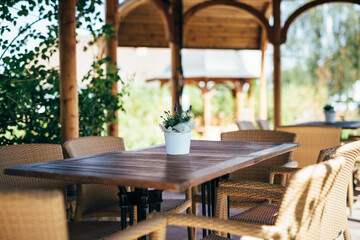 Fototapeta na wymiar Garden sitting in wooden gazebo. In middle is wooden table with metal legs,on which is white flowerpot with decorative fabric and flower. There're 6 knitted chairs around table. Background is blurred.