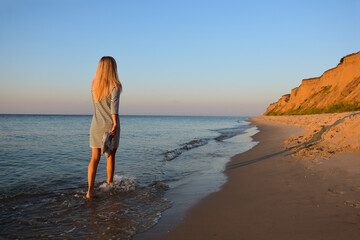 A young blonde girl in the morning goes on the beach barefoot and carries shoes in her hand. Back view