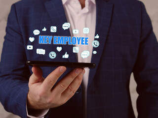  KEY EMPLOYEE inscription on the screen. Budget analyst hands holding black smart phone. A key employee is an employee with major ownership and/or decision-making role in the business