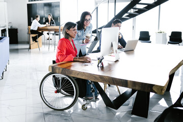 Hispanic transgender woman sitting in wheelchair and coworker looking at computer at workplace in...