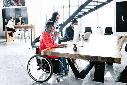 Hispanic Transgender Woman Sitting In Wheelchair And Coworker Looking At Computer At Workplace In Latin America