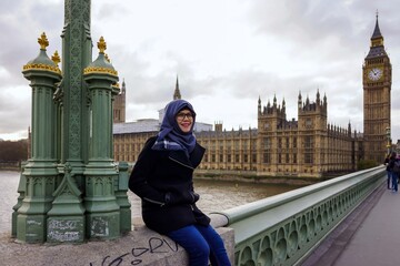 Portrait of beautiful young Asian muslim woman wearing hijab and eyeglasses sitting alone by lamp post on railing of Westminster Bridge. Smiling and happy expression. Westminster palace in background.
