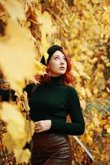 Beautiful young red-haired woman in autumn park. Stylish model among yellow dry foliage of trees. Awesome female in black beret, knitted sweater looks up on sky. Walk in fall nature.