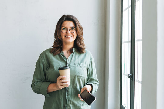 Portrait of Young smiling brunette woman plus size near window with paper cup of coffee and smartphone in hands in bright modern office